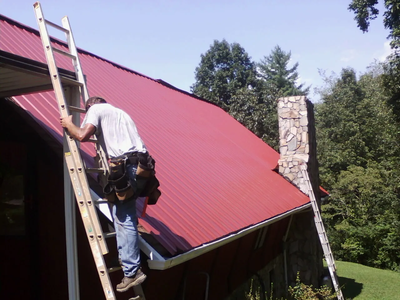 Roofer on a ladder beside a red metal roof and stone chimney — Soffit & Fascia Repair by Roofing Brothers in Green